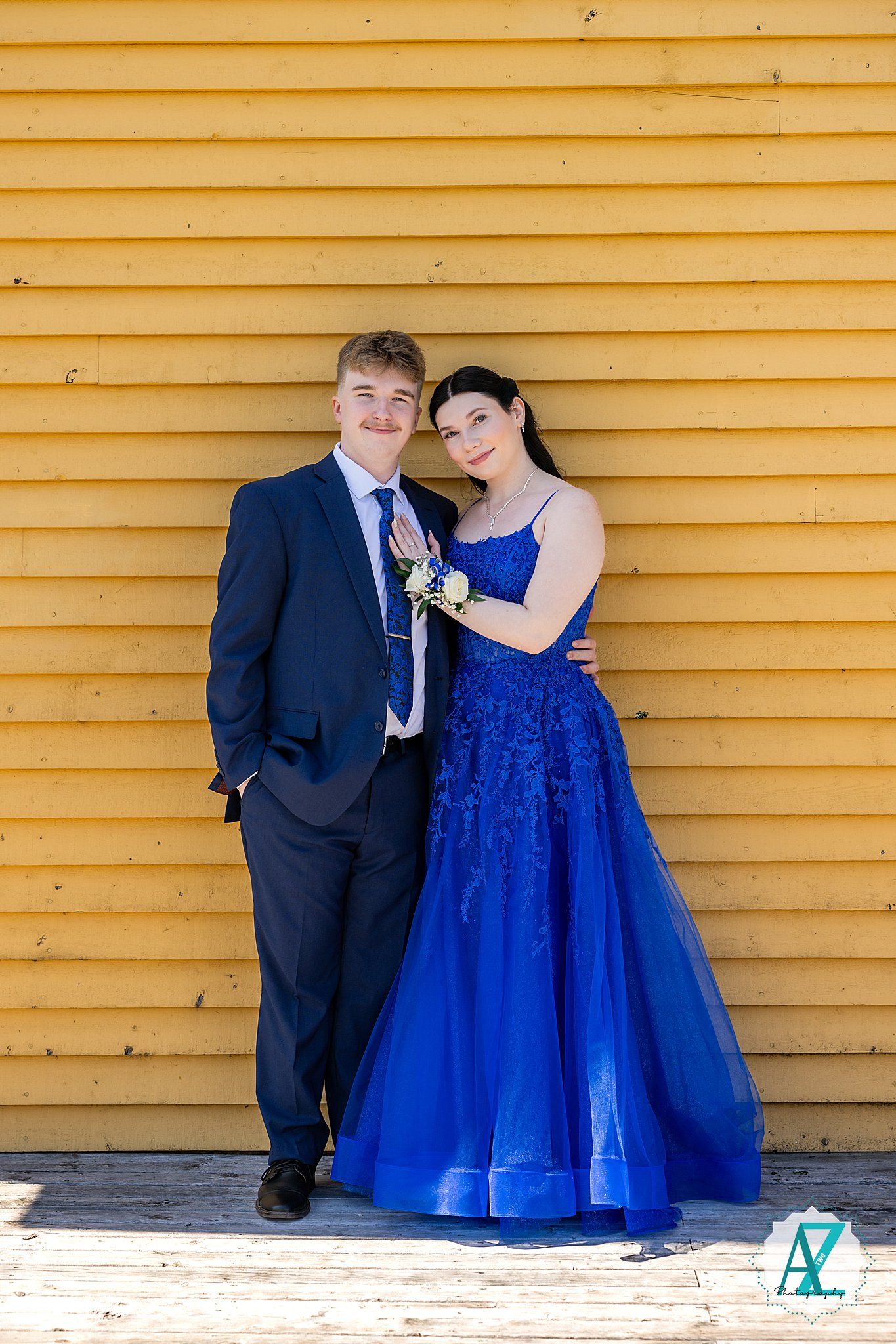 A couple poses together for a formal photo against a yellow wooden wall, with one wearing a blue gown and the other in a navy suit.