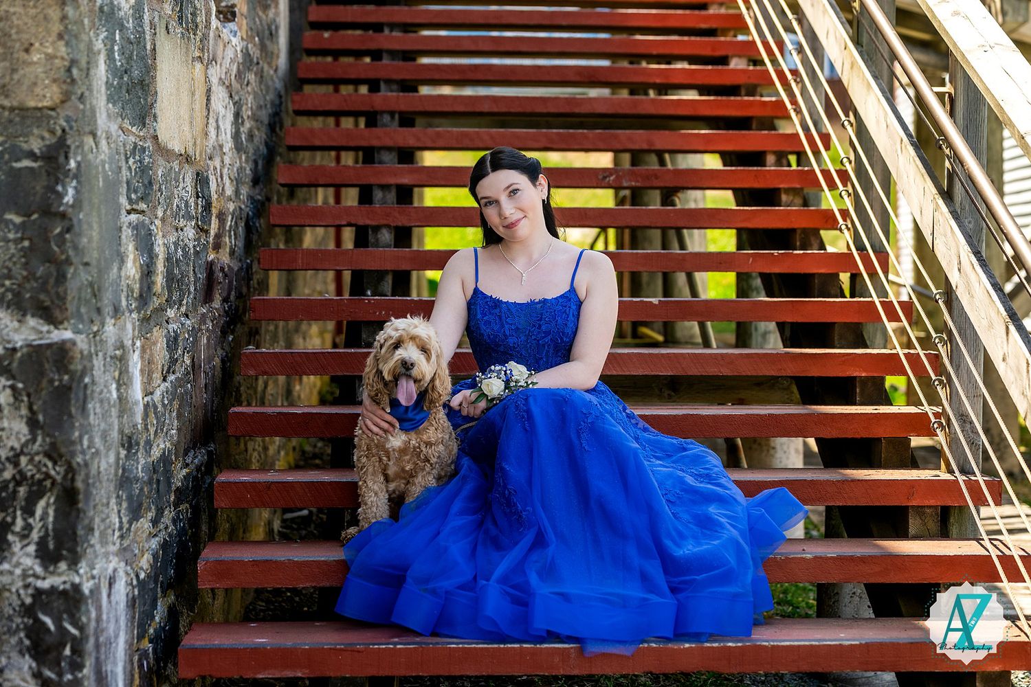 A dog and someone in a flowing royal blue gown sit together on wooden stairs outdoors.