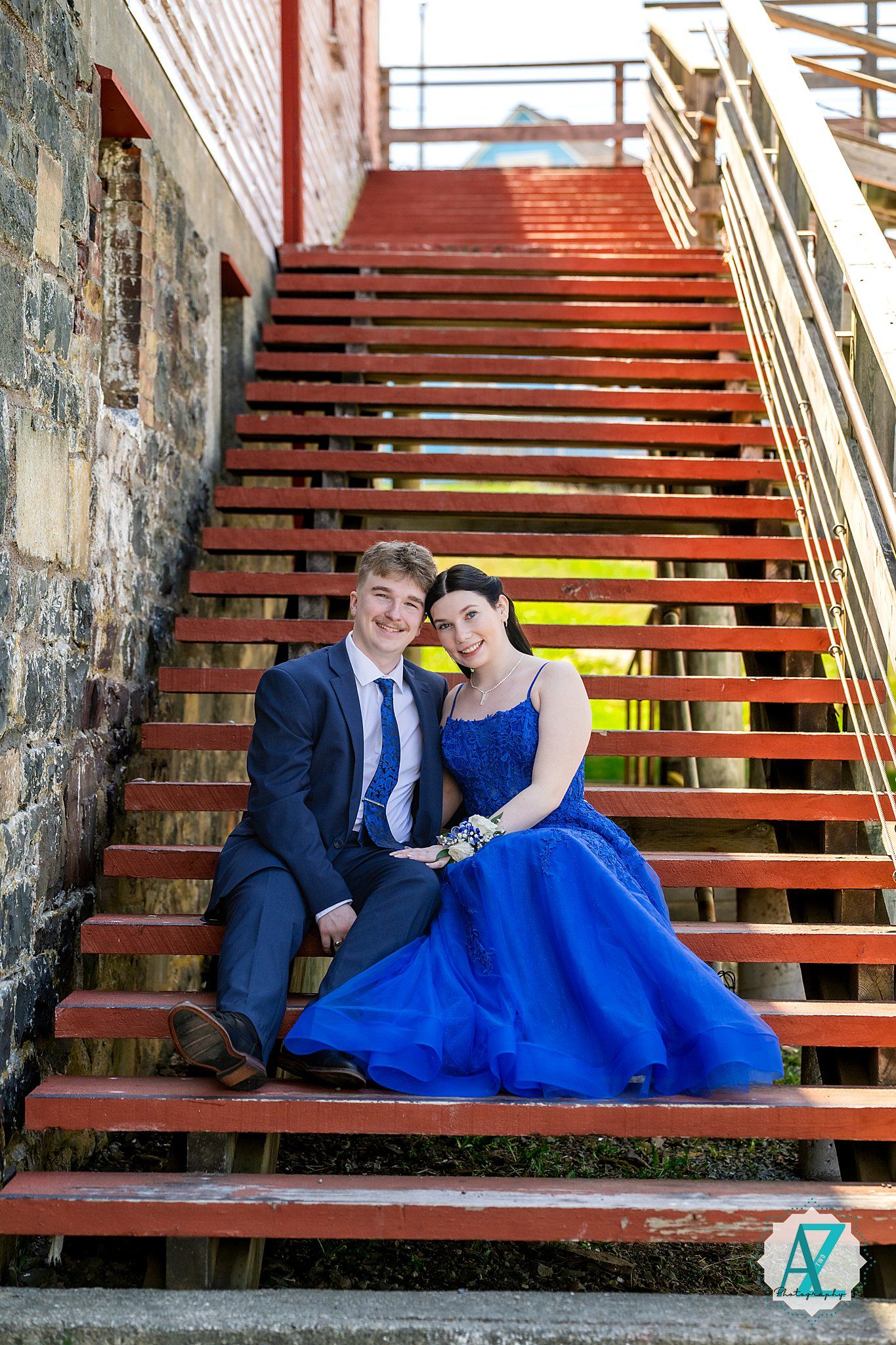 Couple in formal attire sits together on red wooden stairs, with person in vibrant blue gown and dark suit.