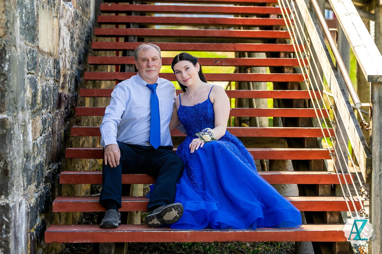 Two people in formal attire sitting together on wooden stairs, one wearing a blue tie and one in a royal blue gown.