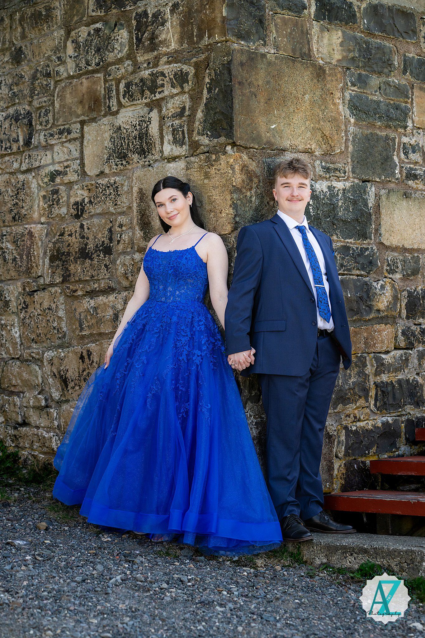 A couple poses together against a stone wall, with one wearing a sparkling blue ball gown and the other in a navy suit.