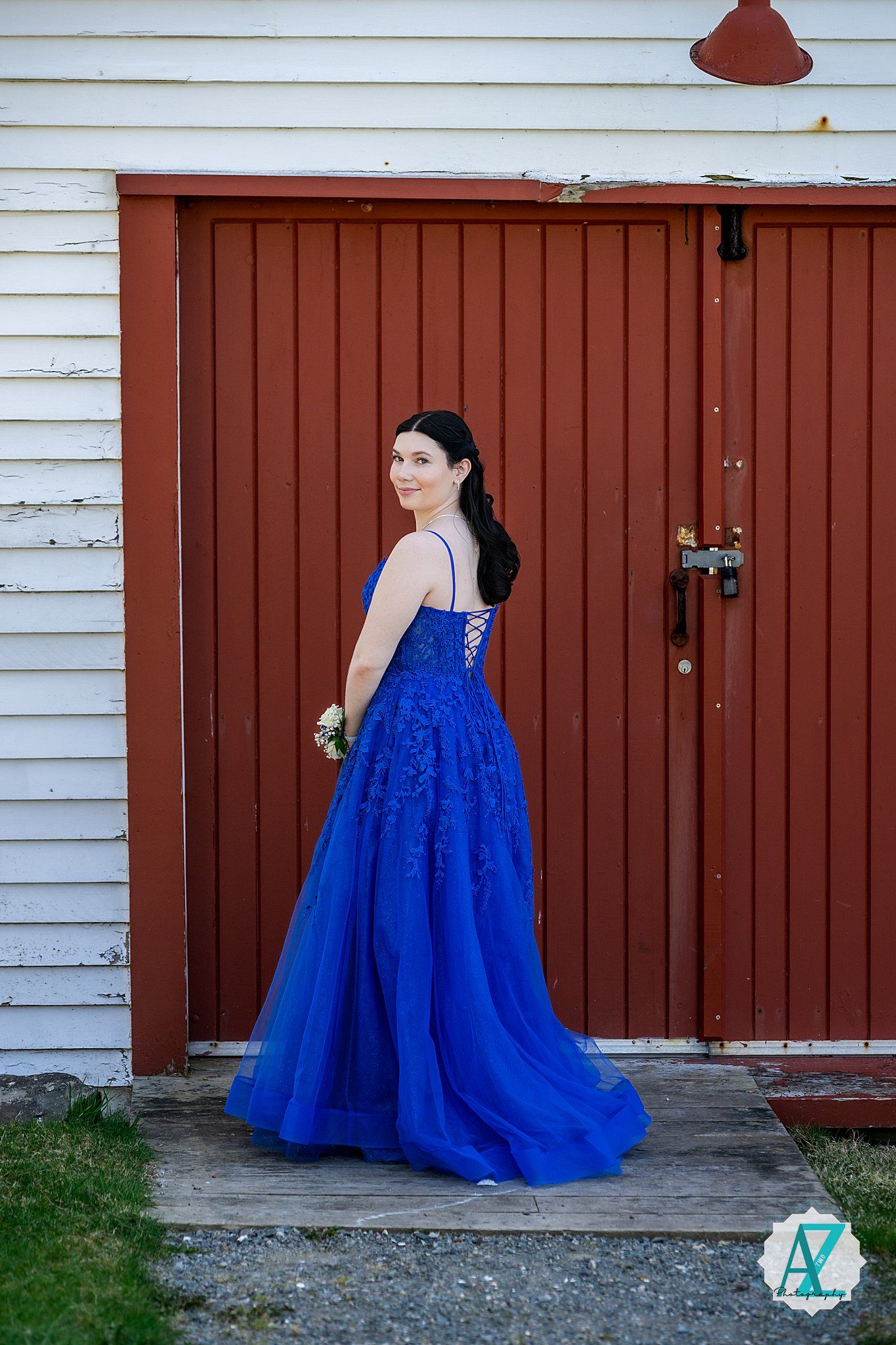 A person in a vibrant royal blue formal gown stands against a rustic red barn door and white siding.
