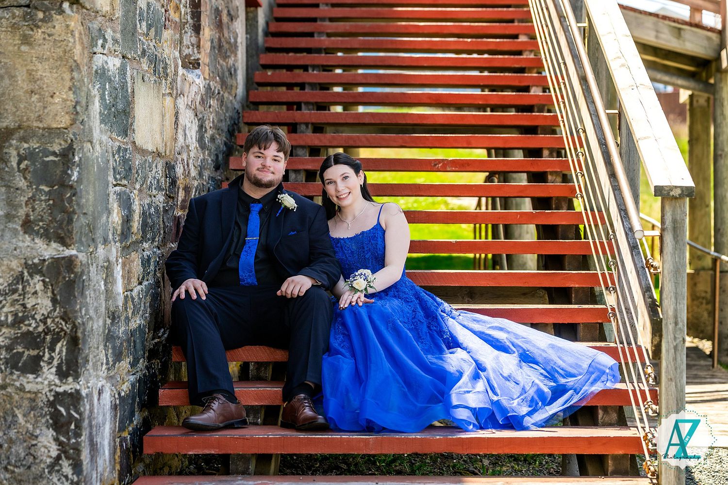 Couple sitting on red wooden stairs with stone wall backdrop as woman's blue gown flows elegantly.