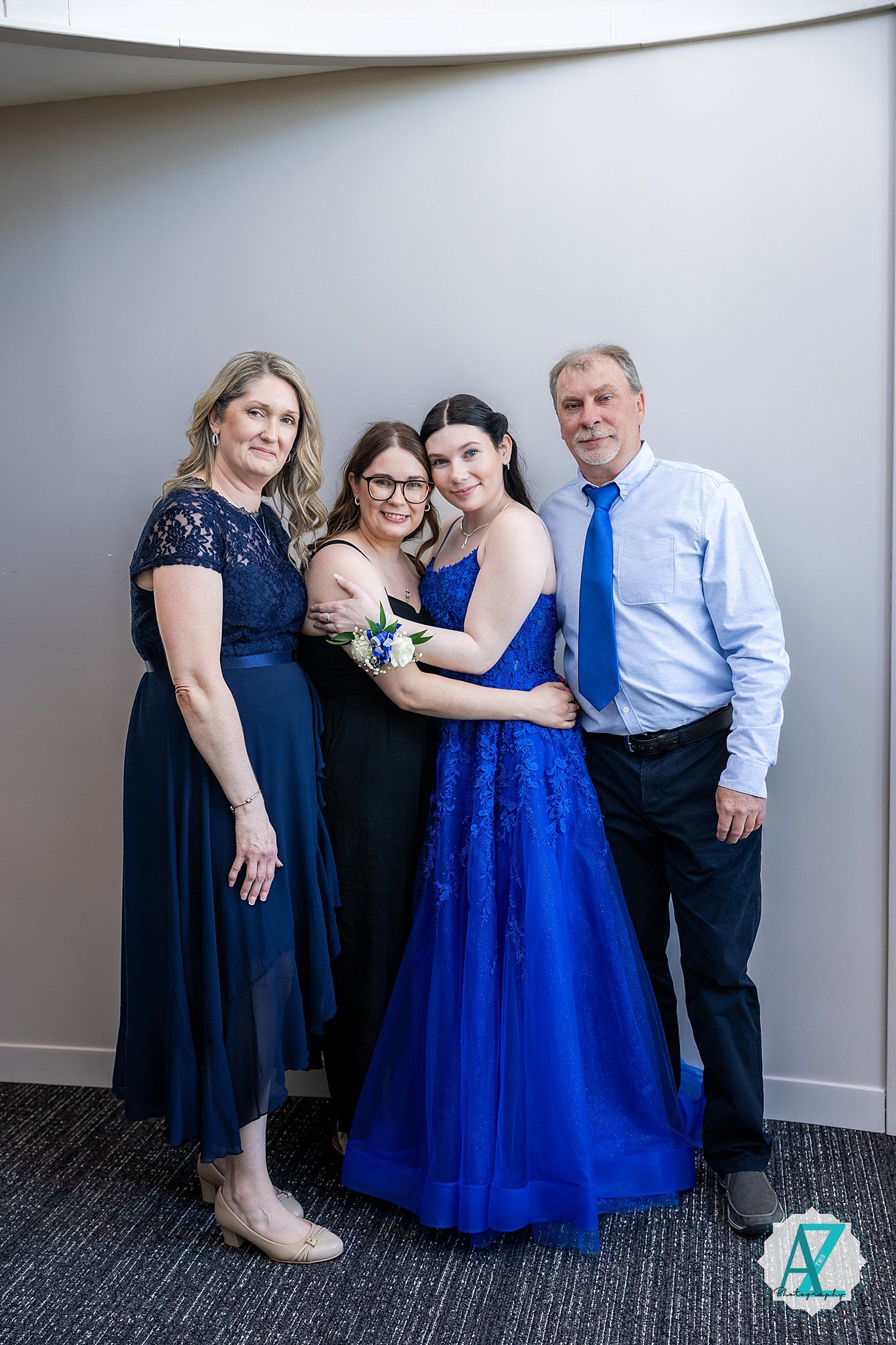 Four people pose together against a light gray wall wearing coordinating navy and royal blue formal attire.