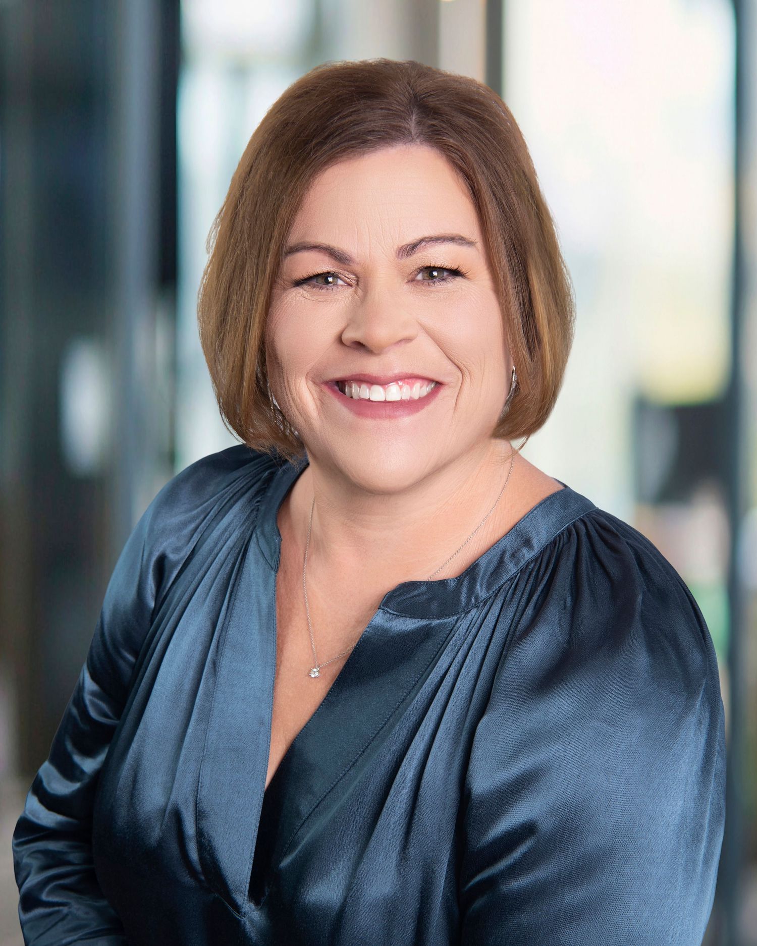 Professional headshot portrait of person wearing navy blue satin blouse against modern office background.