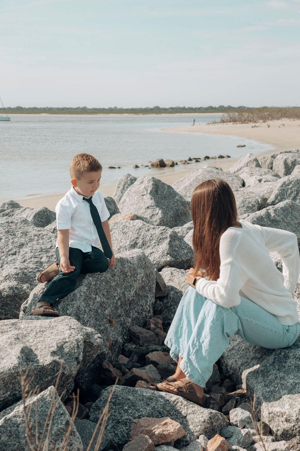 Young boy in white shirt and tie sitting on rocks as woman watches by beach.