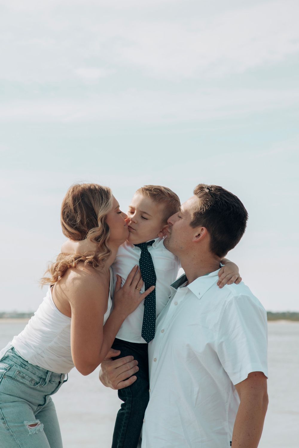 Parents kissing young boy on cheeks simultaneously at beach, child wearing white shirt and tie.