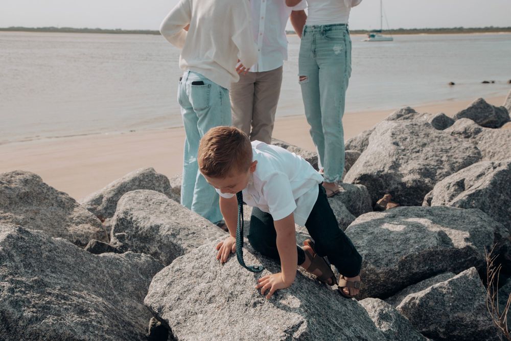 Young boy climbing on rocks at beach while family stands nearby on sunny day.