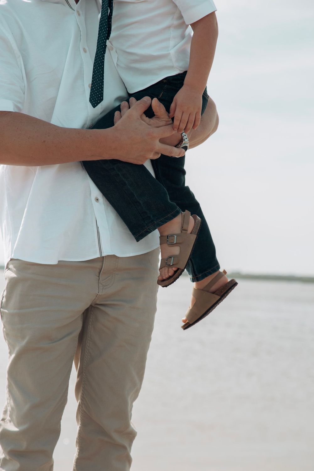 Father carrying young child on beach, wearing white shirt and khaki pants, sandy shore.