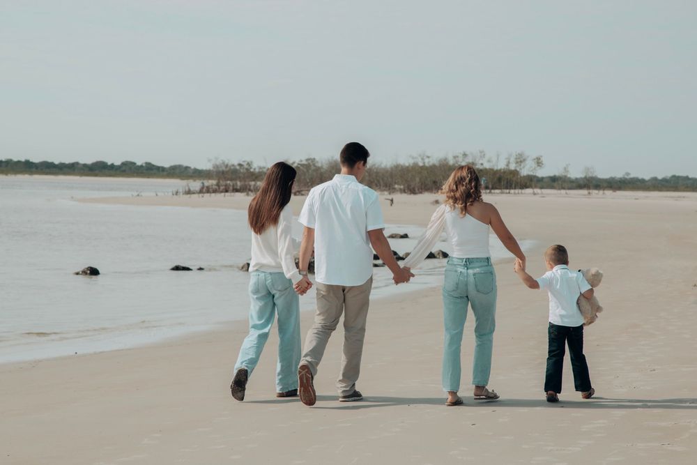 Family of four walking hand-in-hand along sandy beach, wearing coordinated white and blue outfits.