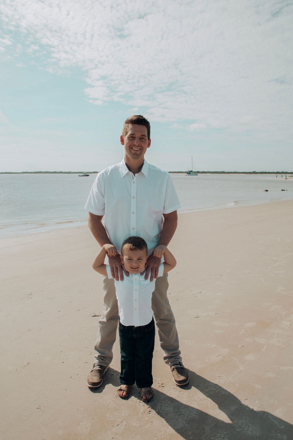 Smiling father standing behind young son on sandy beach with blue sky and water.