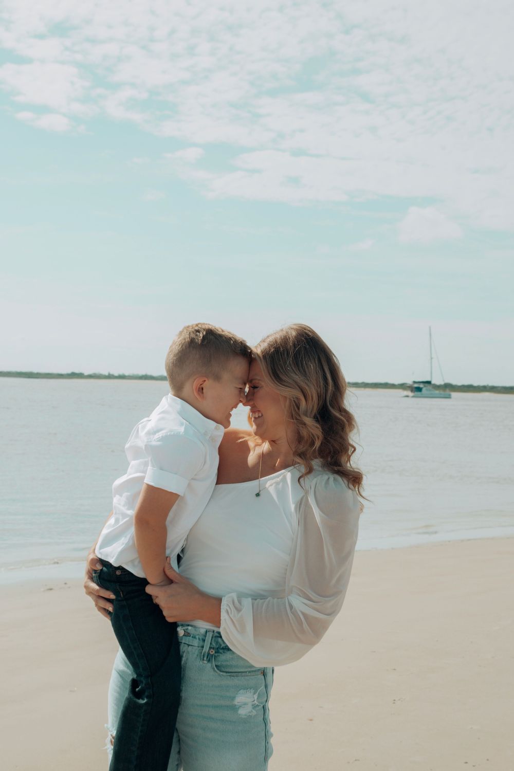 Mother and young son touching foreheads tenderly on a sunny beach with sailboat behind.