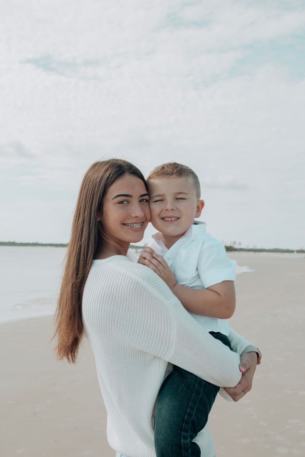Woman in white sweater holding smiling boy on sandy beach with calm water backdrop.