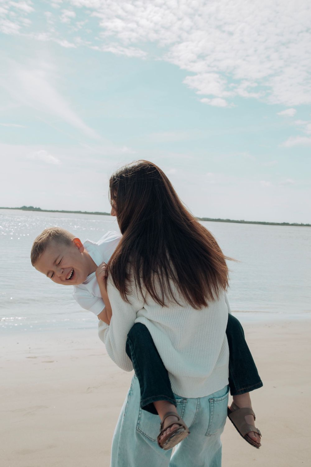 Laughing boy being carried piggyback by dark-haired woman on a bright sandy beach.