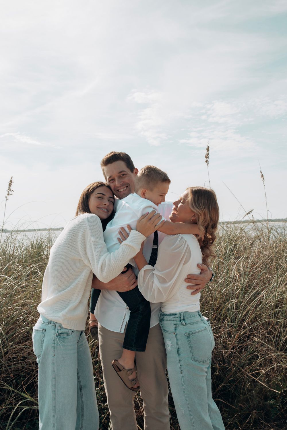 Joyful family embracing in beach grass, parents and two children hugging outdoors.