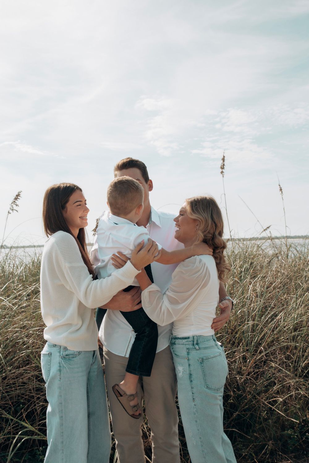 Family of four embracing in tall beach grass, parents holding young child between two siblings.