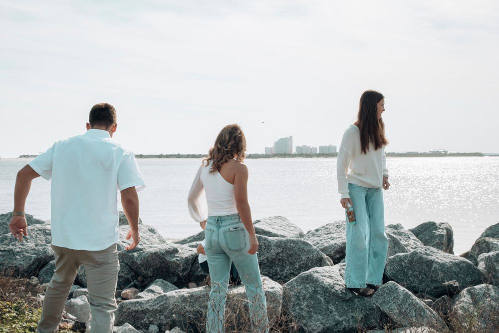Three people standing on rocky shoreline gazing at calm water and city skyline.