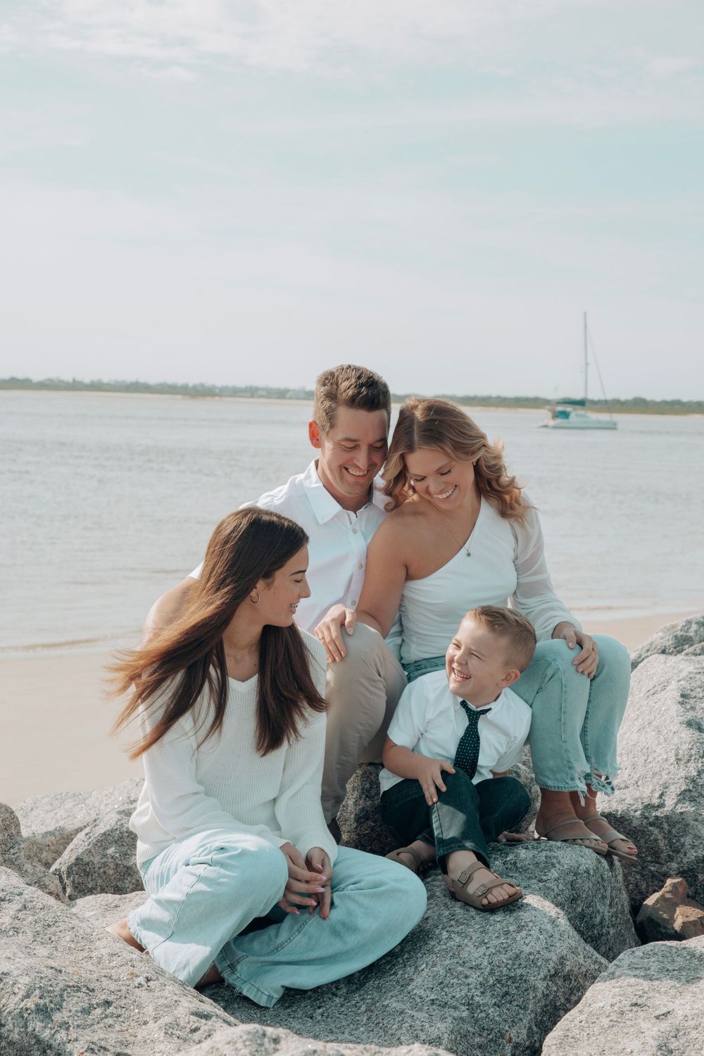 Happy family of four sitting on rocks by the ocean, smiling and laughing together.