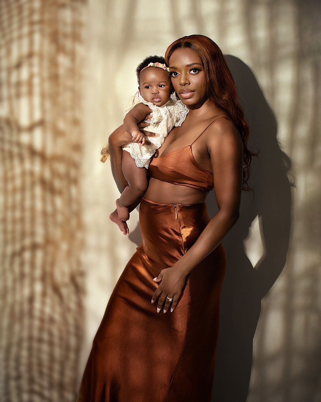 Mother and baby in warm sunlight wearing brown and copper-colored formal attire with shadows casting on wall behind them.