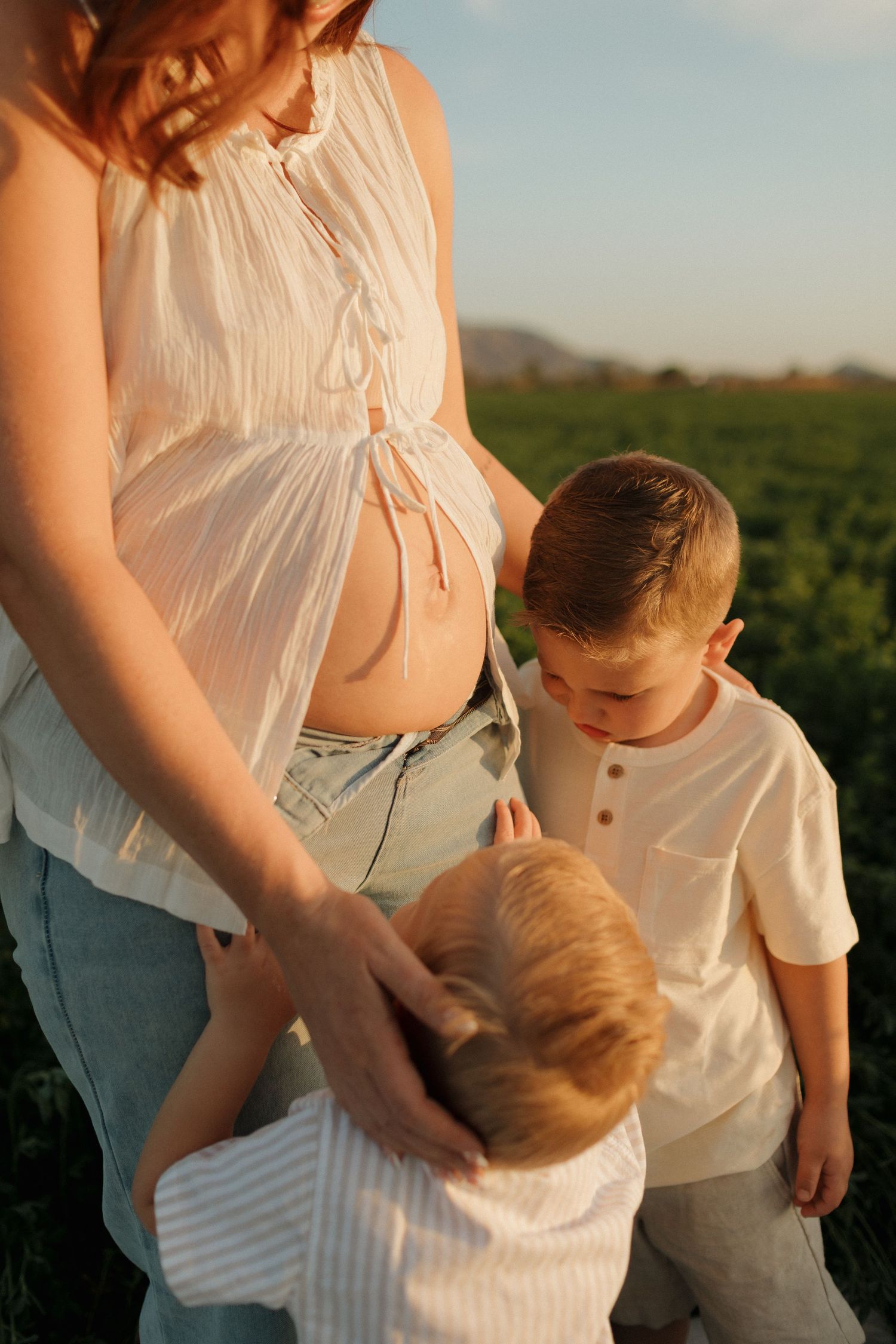 A tender moment in a field at sunset shows a pregnant woman in a white blouse with two young children.