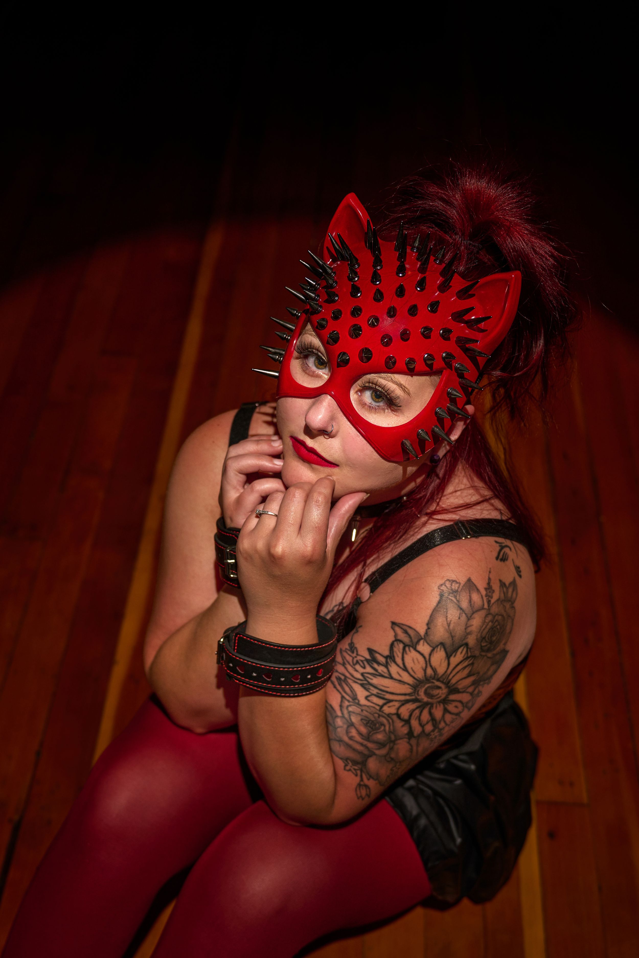 Person wearing spiked red cat mask and dark attire poses dramatically with tattoo visible in moody red lighting.