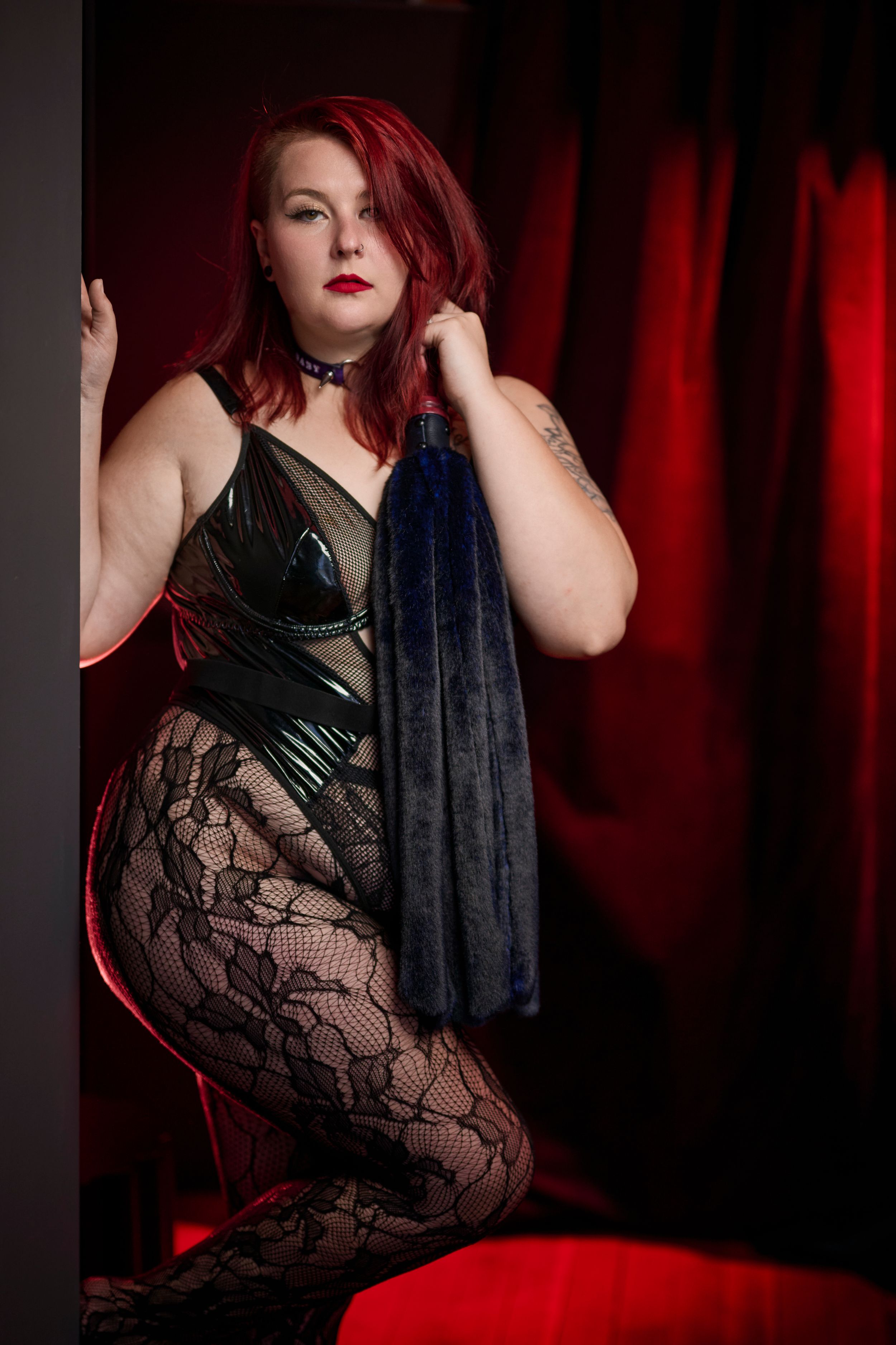 A figure in black lace and leather poses dramatically against red curtains in moody boudoir lighting.