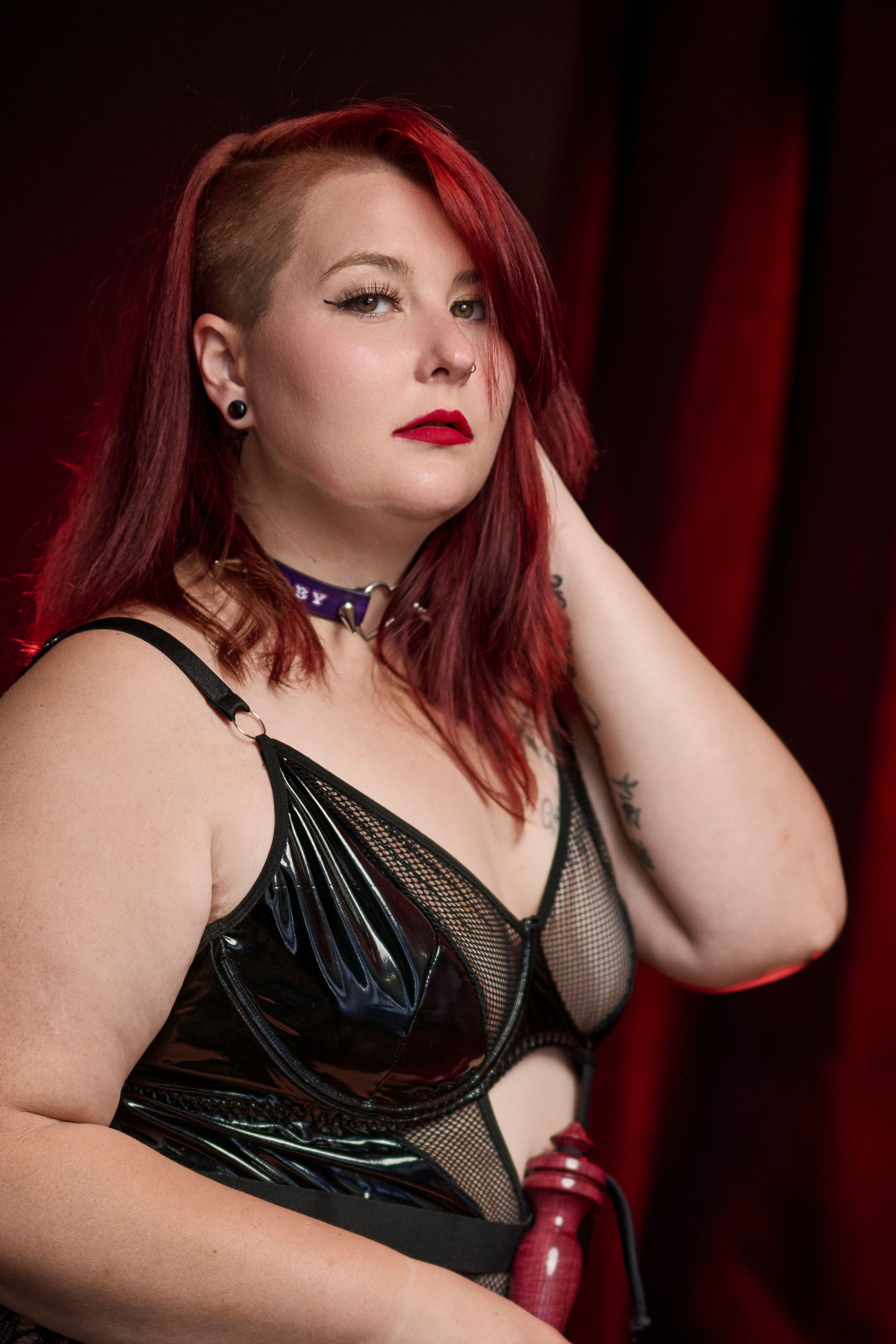 Figure in red hair and black vinyl corset poses against dark red curtain backdrop with dramatic lighting.