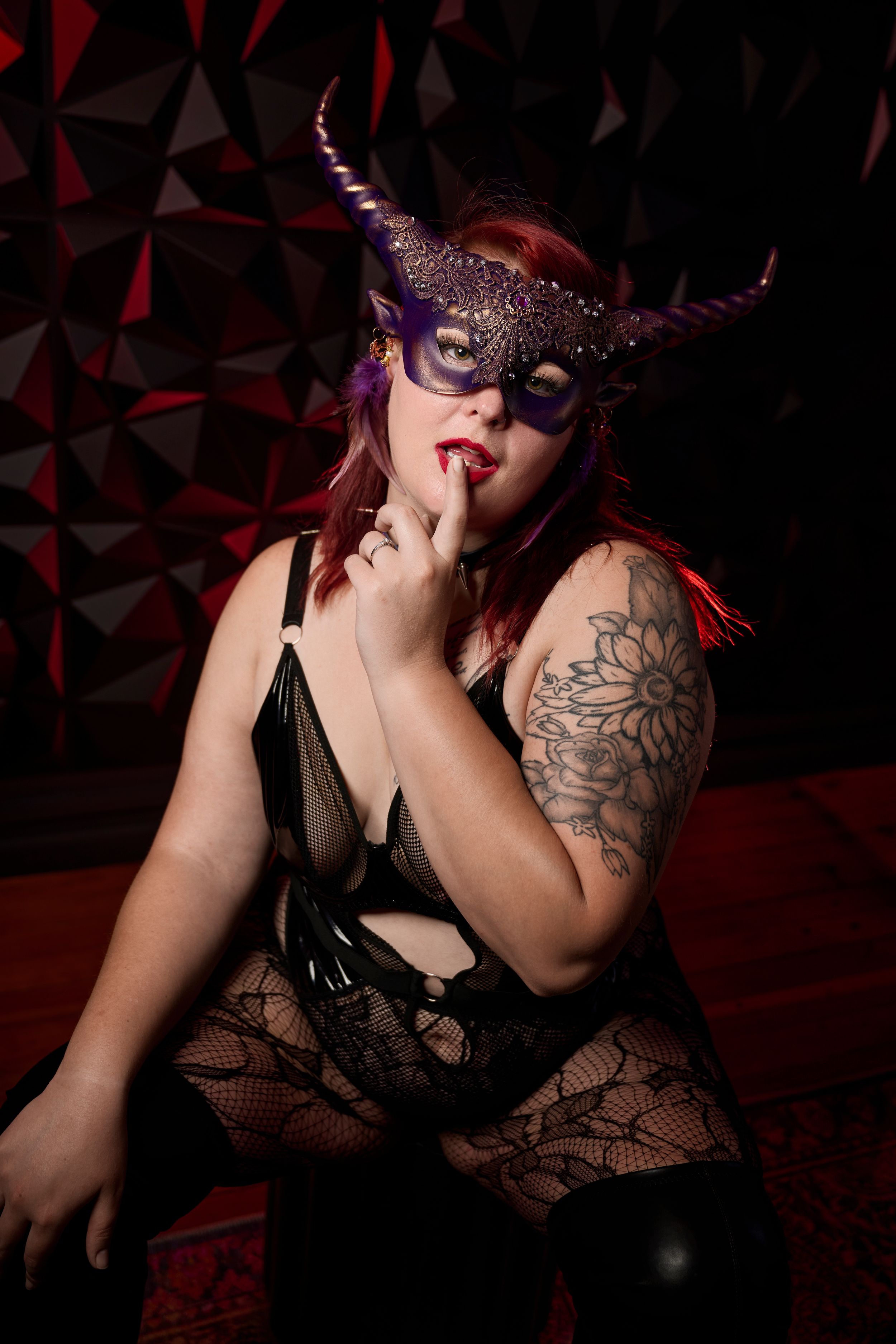 A masked figure in dark attire poses mysteriously against a geometric red backdrop with a finger to their lips.