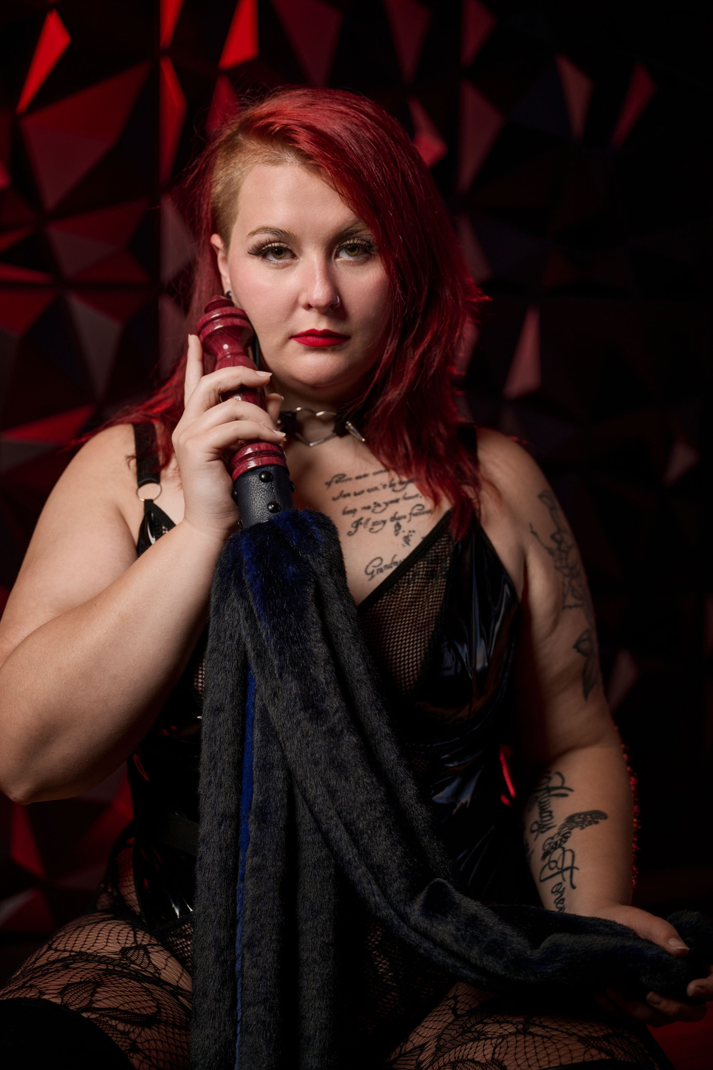 A tattooed singer in Gothic attire poses with a microphone against a dramatic red geometric backdrop.
