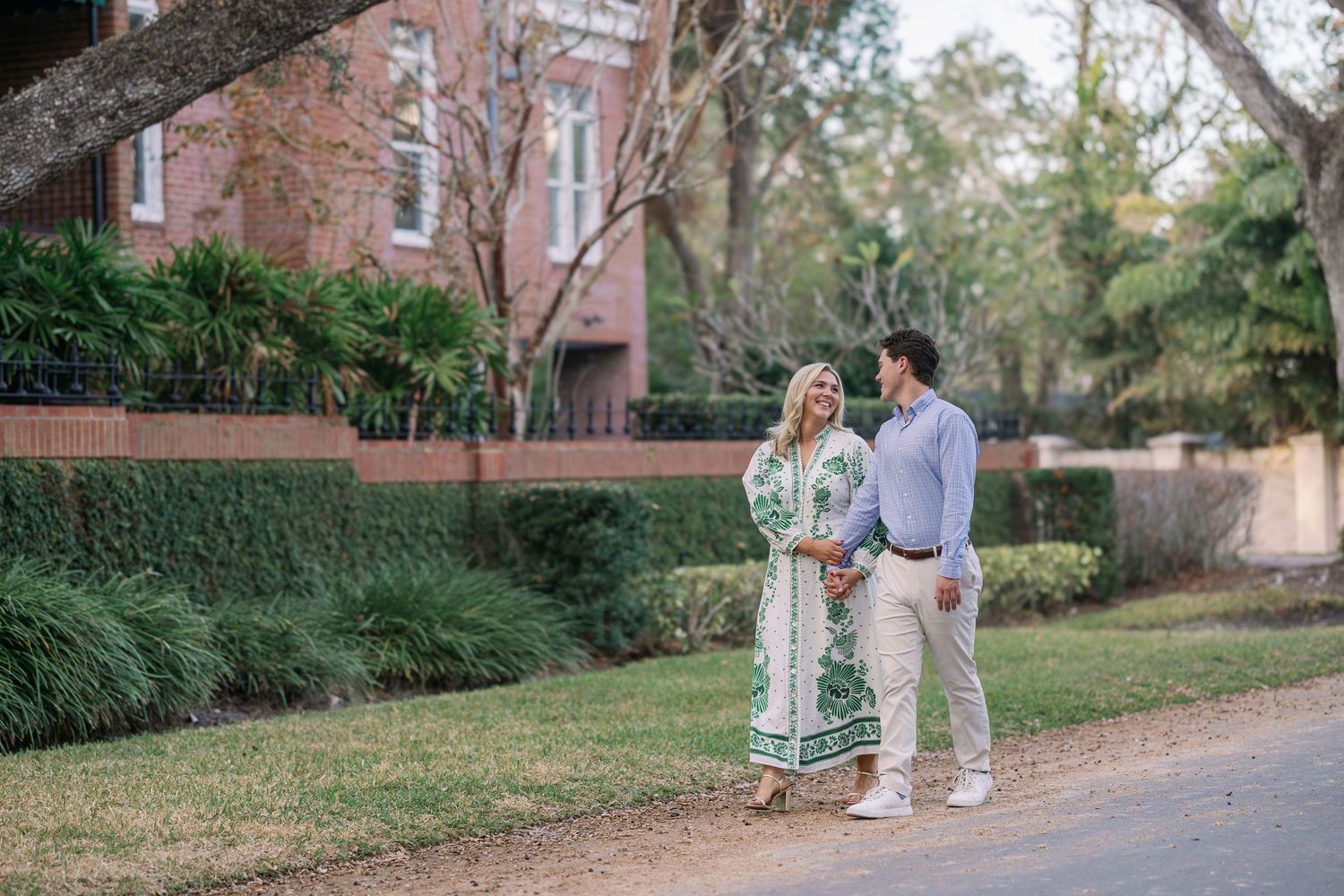A couple standing together on a path beside a brick building and lush garden on a sunny afternoon.