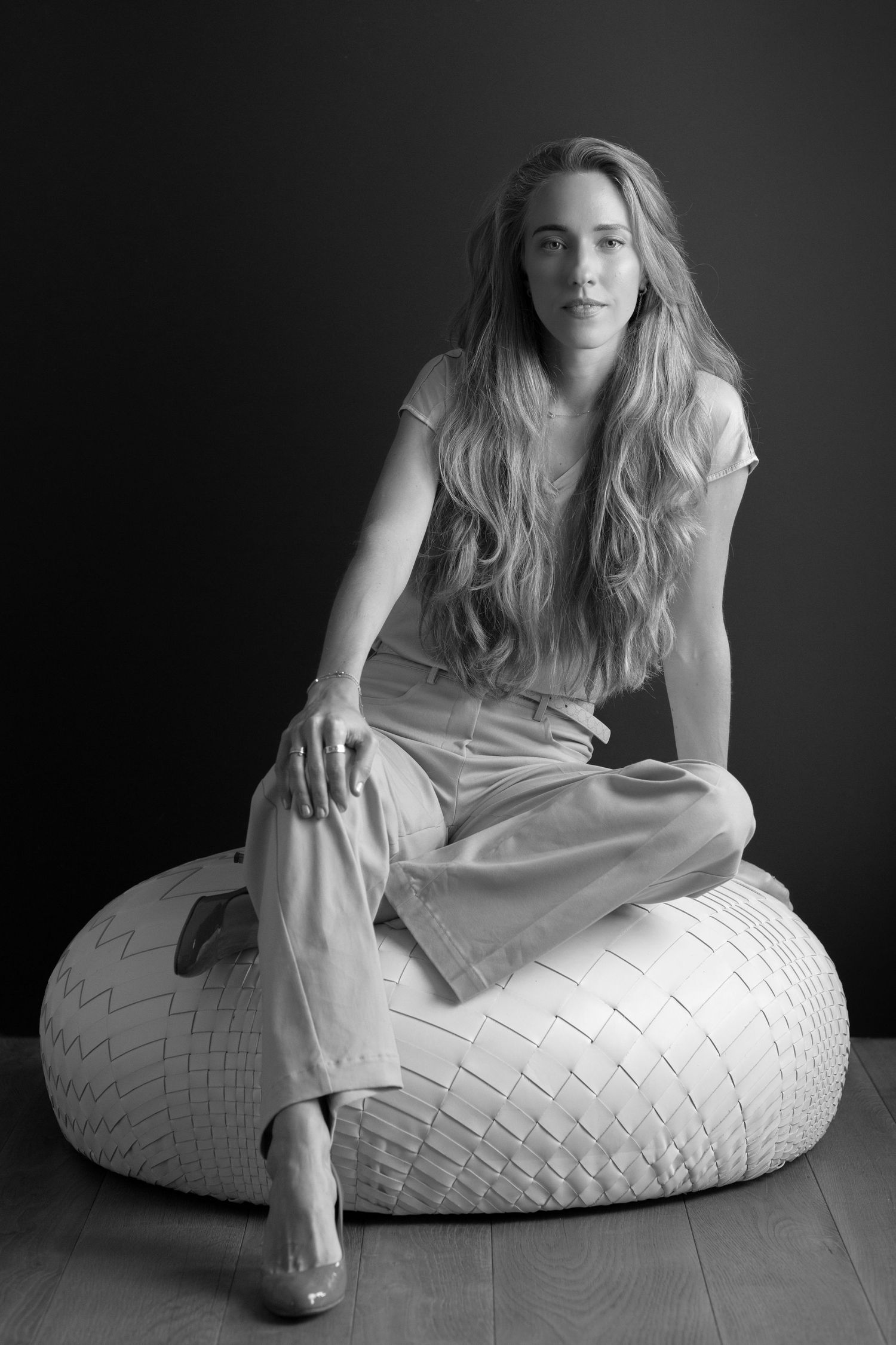 Artistic black and white portrait of a person with long wavy hair sitting casually on a woven ottoman pouf in a minimalist setting.