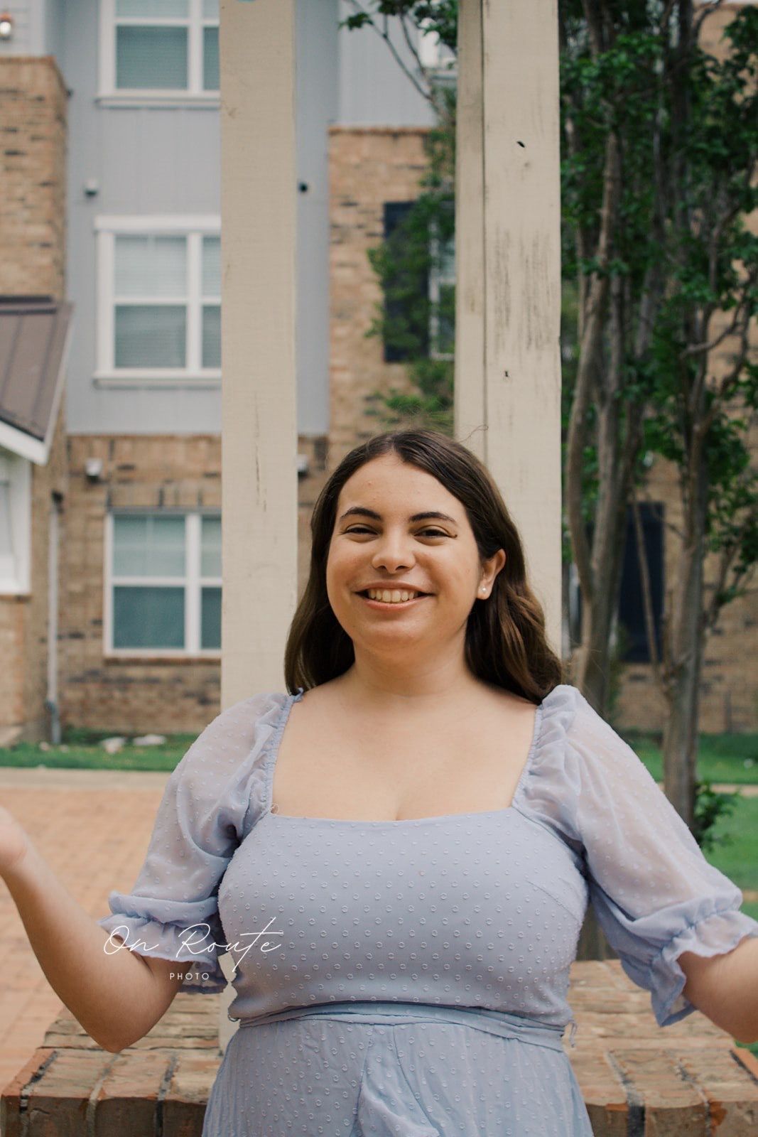 Portrait photo outdoors showing someone in a light purple dress with puff sleeves against a brick and stone building background.