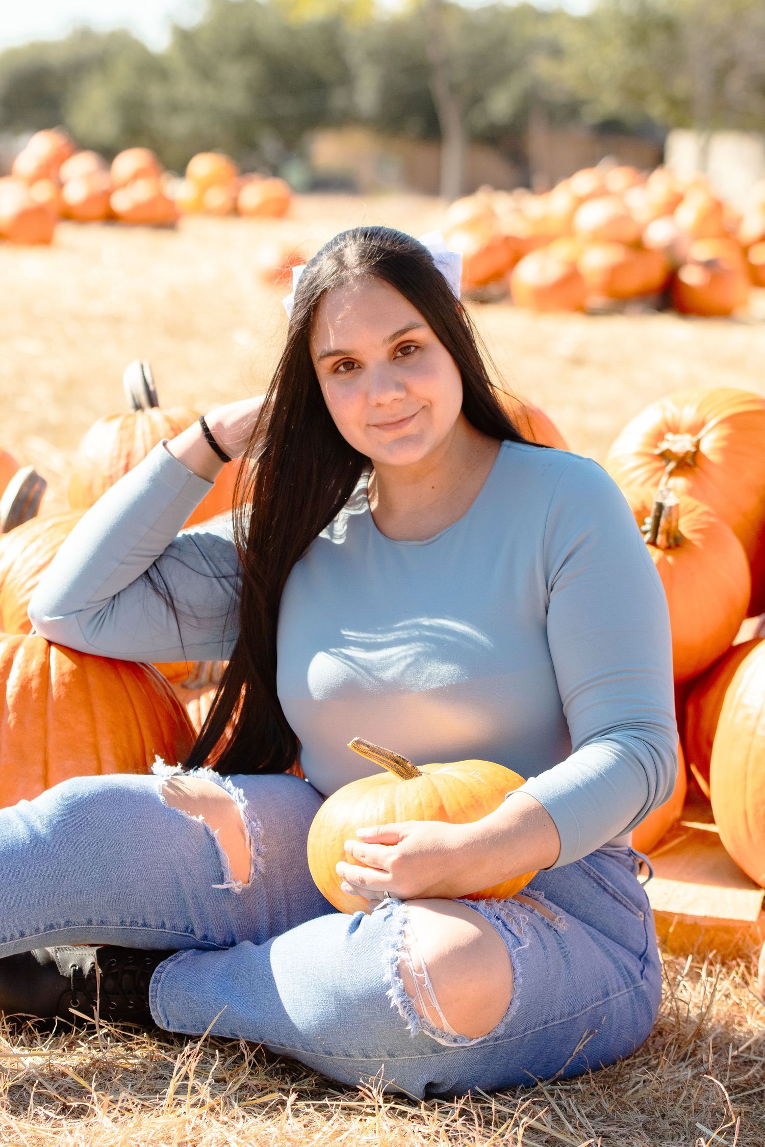 Autumn scene at pumpkin patch with someone sitting among orange pumpkins wearing light blue top and ripped jeans.