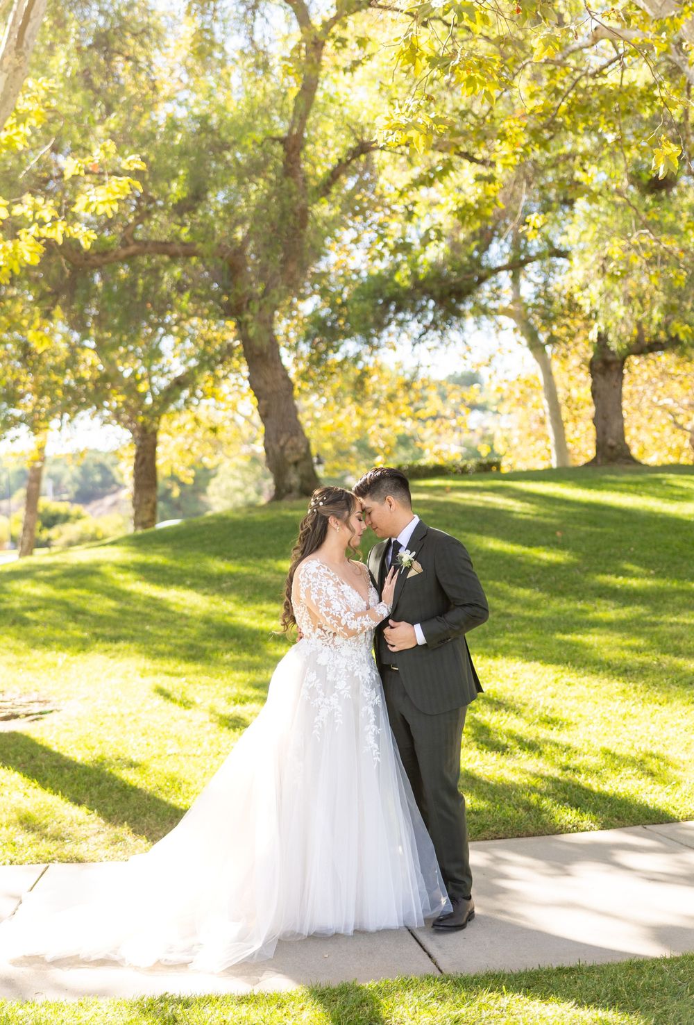 A romantic wedding couple shares an intimate moment on a sunlit hillside beneath autumn trees.