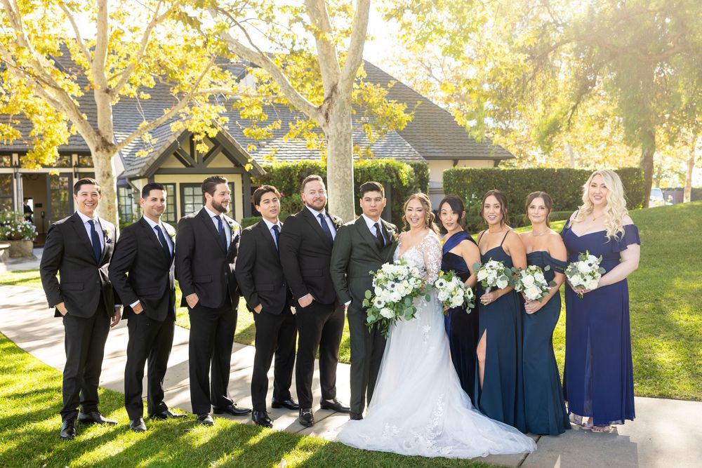 A wedding party in formal attire poses together on a sunny lawn with a building in the background and autumn trees.