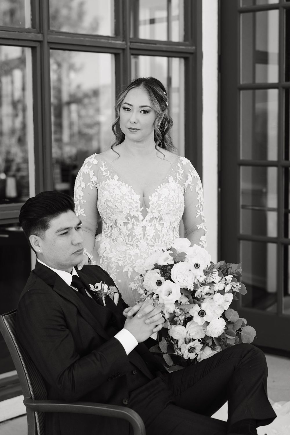 Elegant bride in lace gown stands beside seated groom holding white floral bouquet in black and white photo.