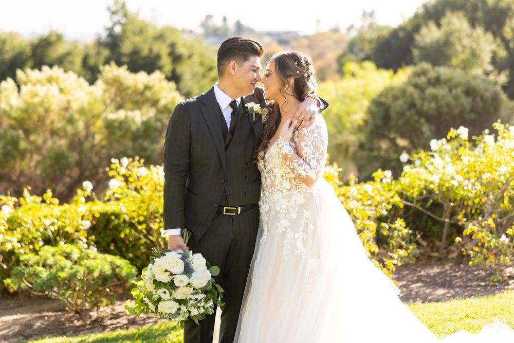 Couple shares intimate moment in sunlit garden with bride in detailed lace gown and groom in dark suit.