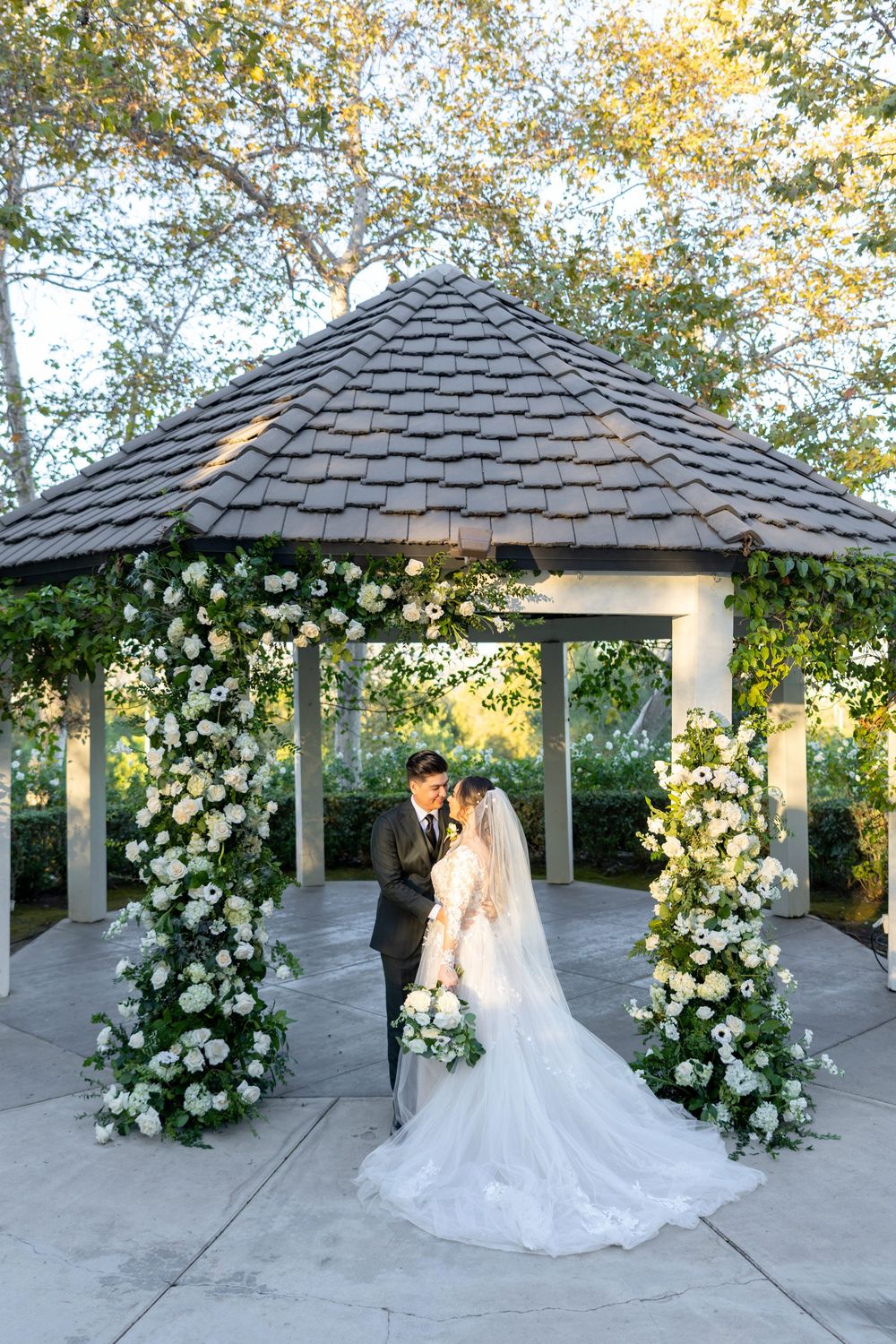 A romantic wedding portrait under a gazebo adorned with white floral arrangements in a garden setting.