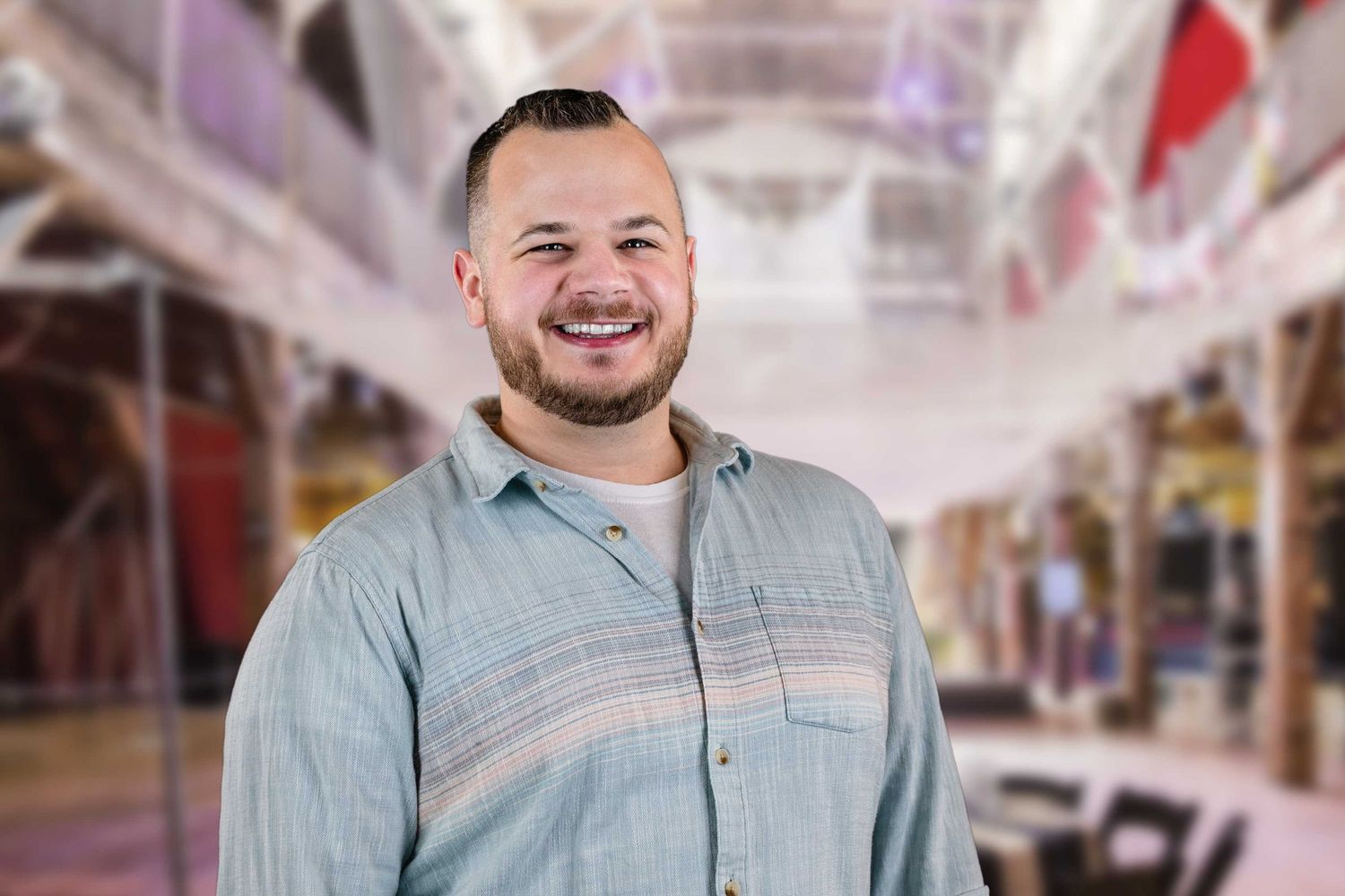A smiling person wearing a light blue button-up shirt stands in a bright indoor venue with hanging decorations.