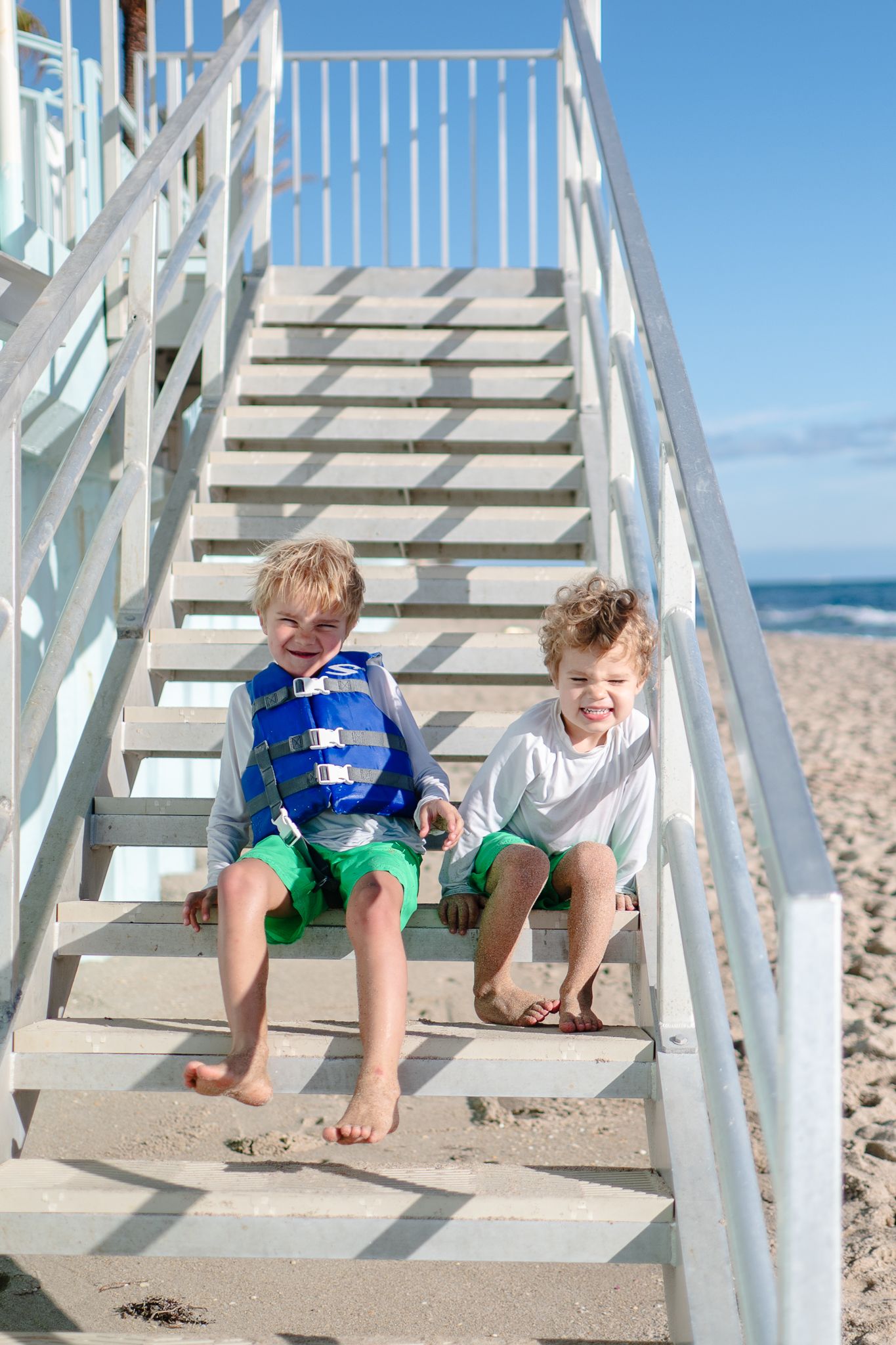 Two children sit together on white wooden stairs at a sunny beach while smiling and wearing casual summer attire.