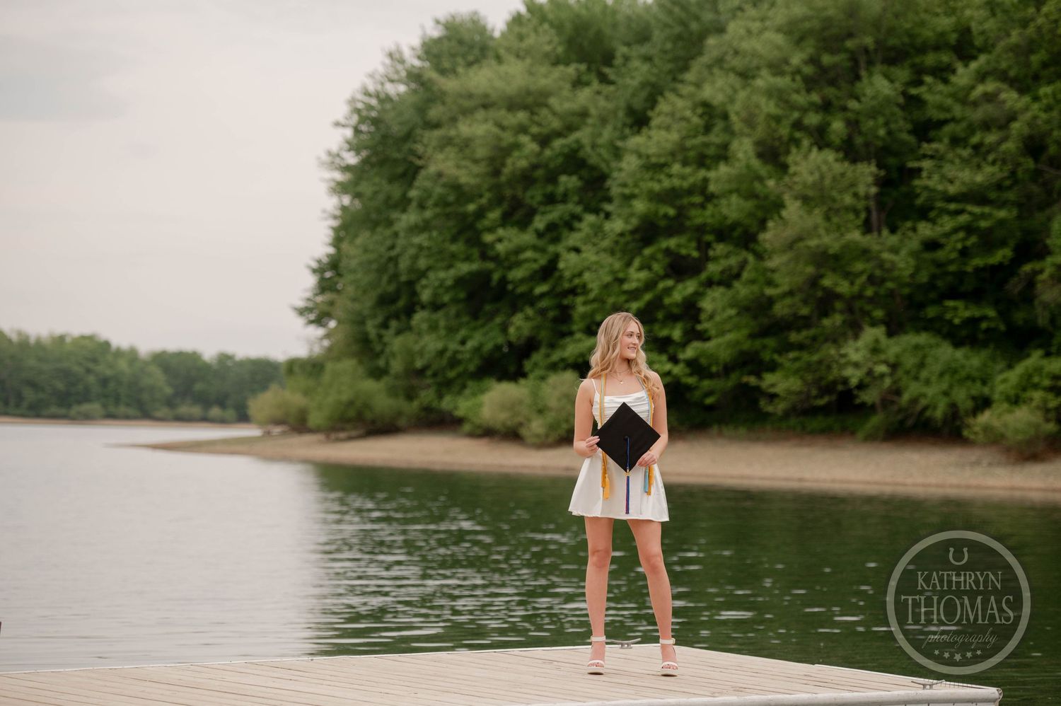 A person in a white dress stands on a wooden dock overlooking a tranquil lake surrounded by green forest.