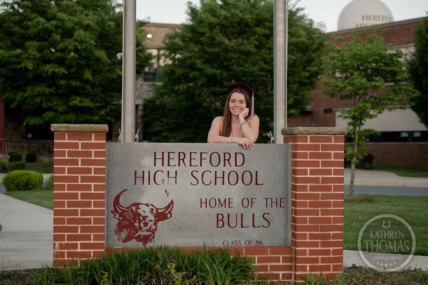 A graduate poses at the brick entrance sign of Hereford High School with its bull mascot logo and trees in background.