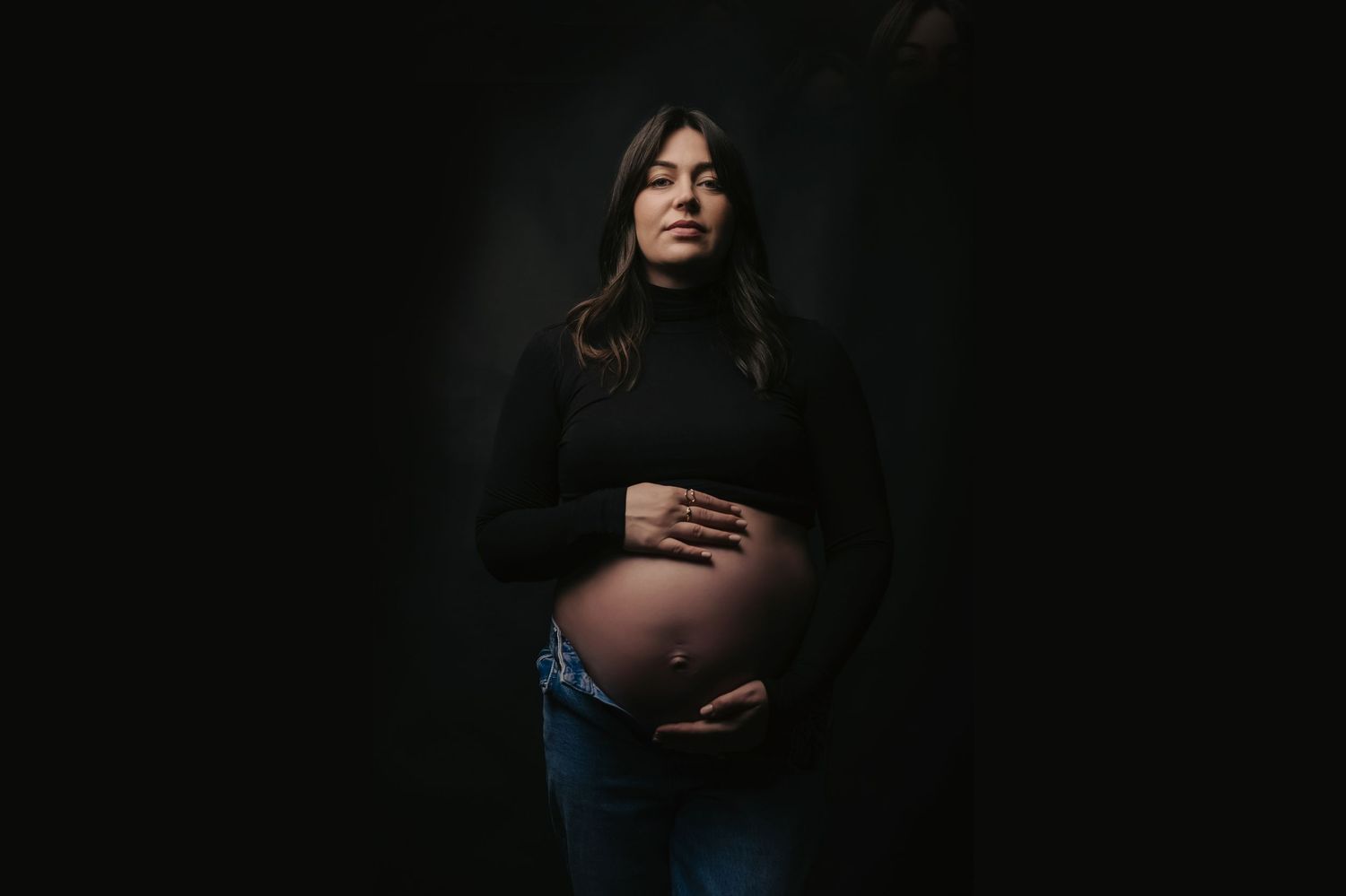 A moody maternity portrait in dark tones shows an expectant mother in black turtleneck and jeans against dark background.