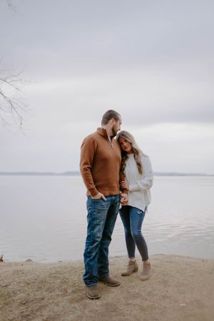 Couple stands close together on a beach with cloudy skies.