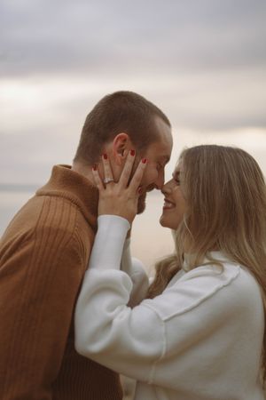 Romantic couple in brown and white sweaters sharing an intimate moment against a cloudy sky.