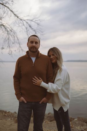Couple in casual sweaters posing playfully by waterfront on cloudy day.