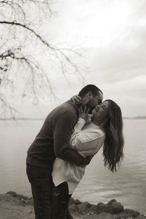 A romantic couple embraces and kisses by a misty waterfront on an overcast day.