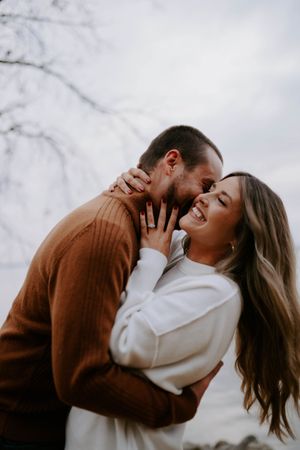 A couple shares an intimate moment wearing brown and white sweaters against a cloudy sky.