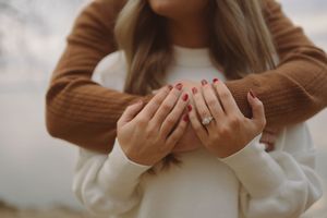Detail shot of hands with red nails and engagement ring against neutral clothing.