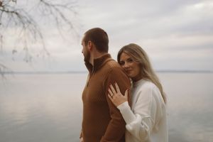 A couple embraces by the water on an overcast day wearing brown and white sweaters.